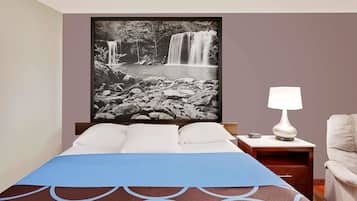 Una cama bien tendida con colcha de tonos azules y marrones, un almohadón blanco y una lámpara blanca sobre una mesita de noche de madera. En la pared, encima de la cama, hay una fotografía en blanco y negro de una cascada enmarcada.