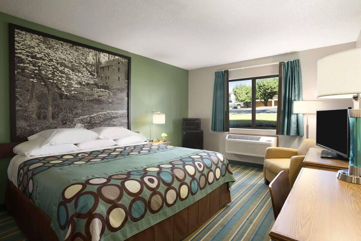 A hotel room with a bed, a desk, a TV, a window with curtains, and a painting on the wall.