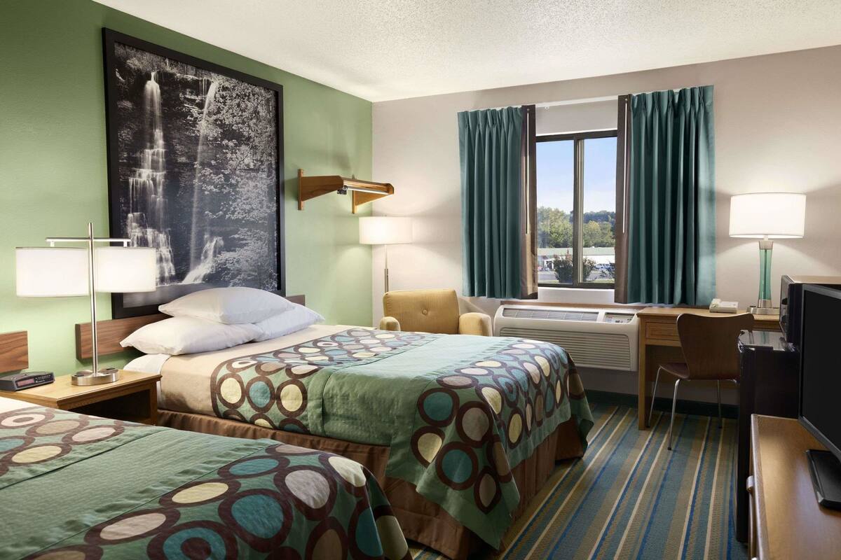 A hotel room with two beds, a desk, a chair, a TV, a lamp, a window with curtains, and a large wall painting.