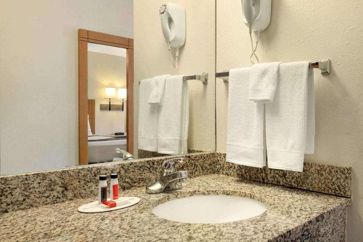 A bathroom with a granite countertop, a sink, a mirror, two towel racks, and a hairdryer mounted on the wall.