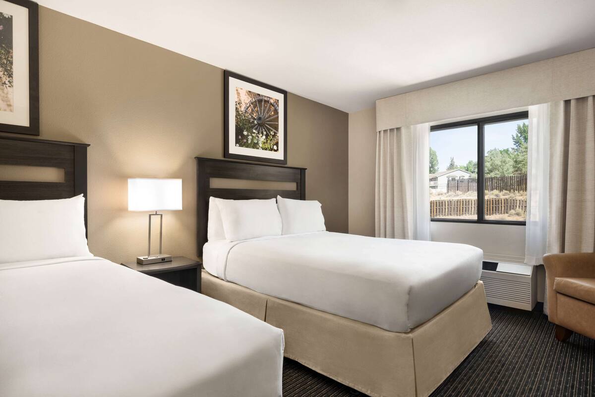 A hotel room with two beds, a nightstand with a lamp, a chair, and a large window with a view of the outdoors.