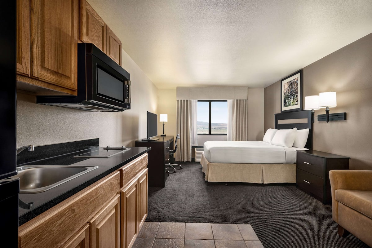 A hotel room with a kitchenette, a bed, a desk, and a window with curtains.
