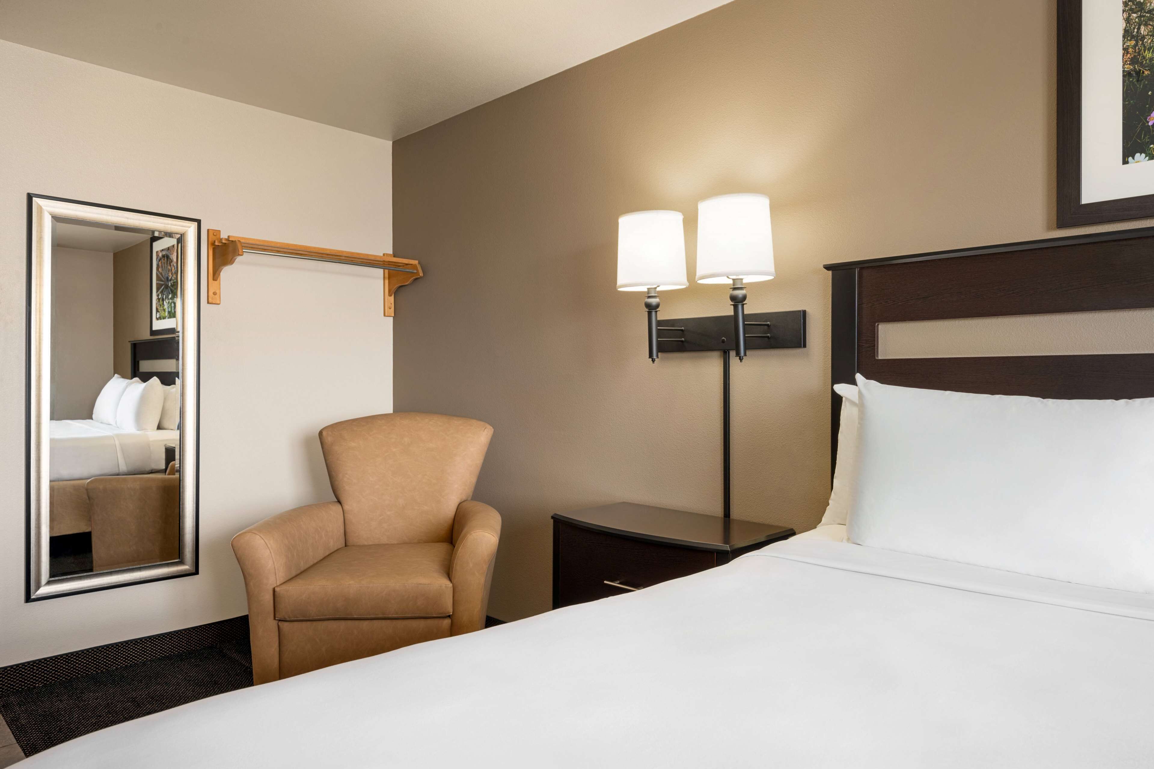 A hotel room with a bed, a chair, a nightstand, a mirror, and a wall-mounted lamp.
