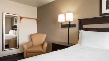 A hotel room with a bed, a chair, a nightstand, a mirror, and a wall-mounted lamp.
