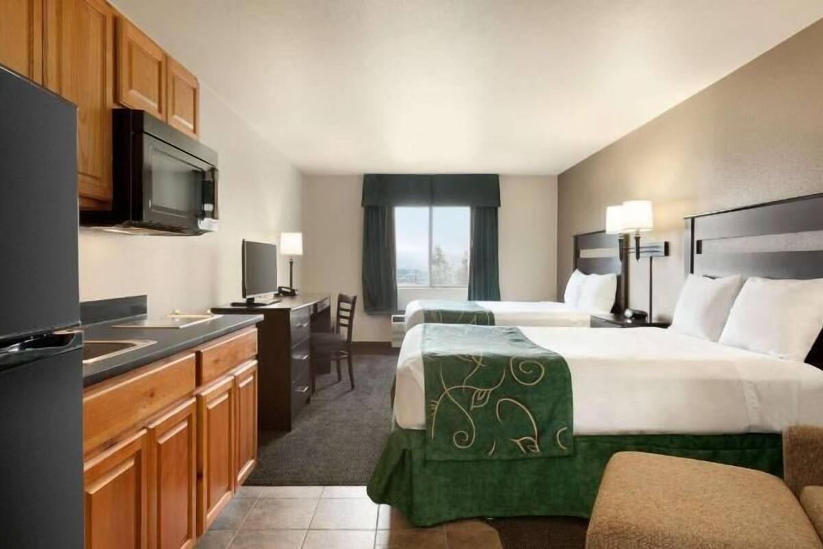 A hotel room with a kitchenette, two beds, a desk, and a view of the outdoors.