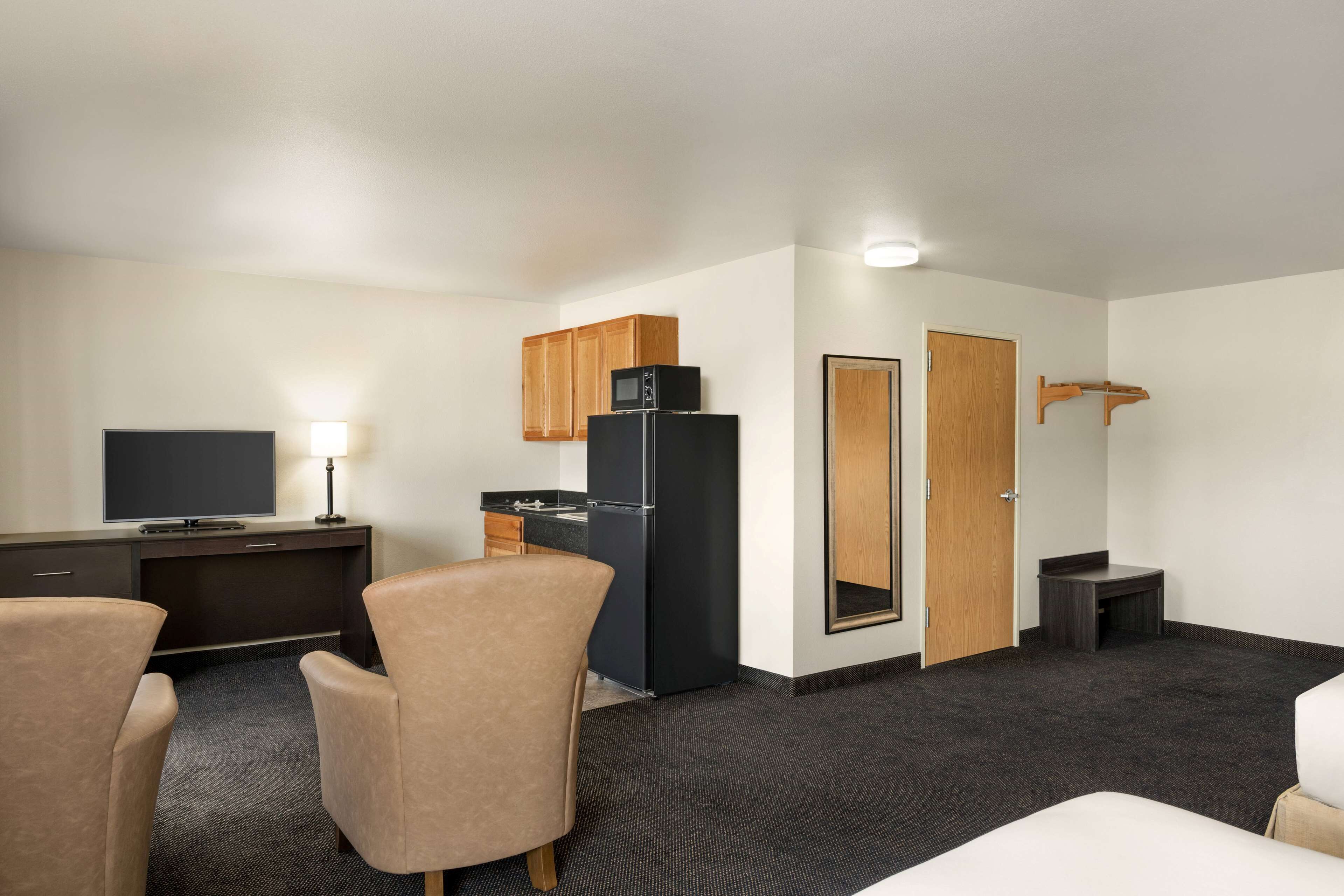 A hotel room with a desk, a black refrigerator, a television, two chairs, a small table, and a door.