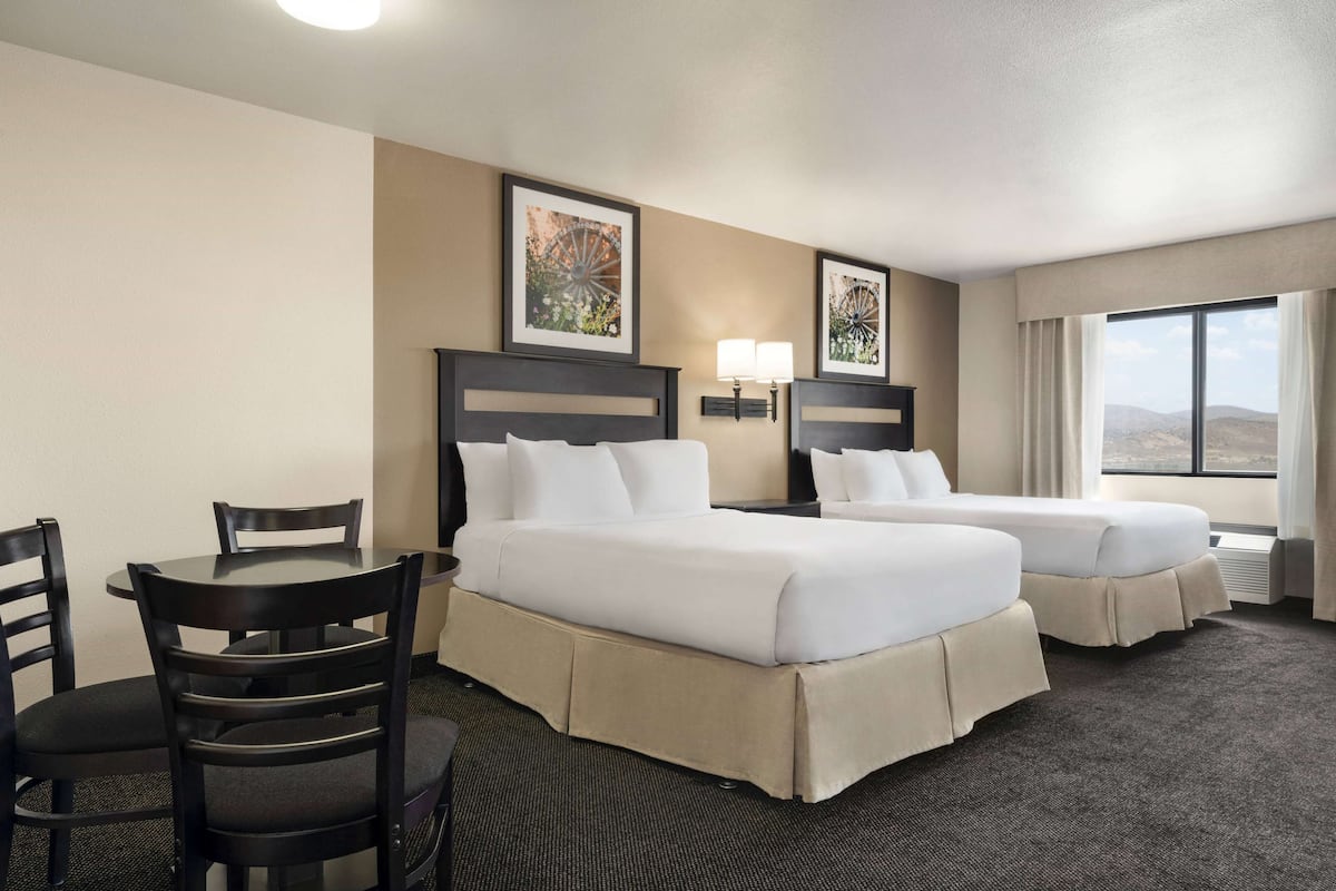 A hotel room with two beds, a dining table with chairs, and a large window offering a view.