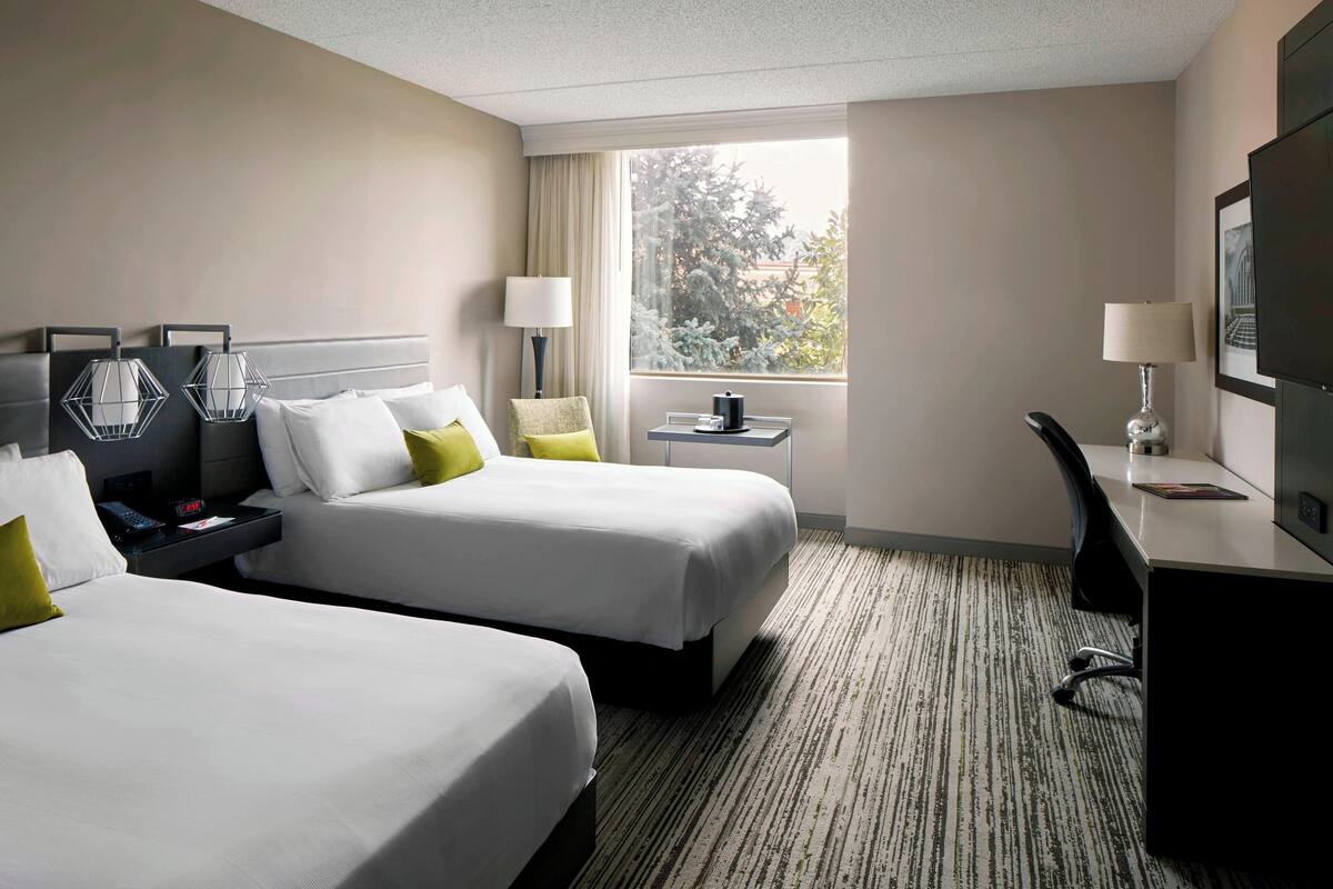 A hotel room with two beds, a desk, a chair, a TV, and a window with a view of trees.