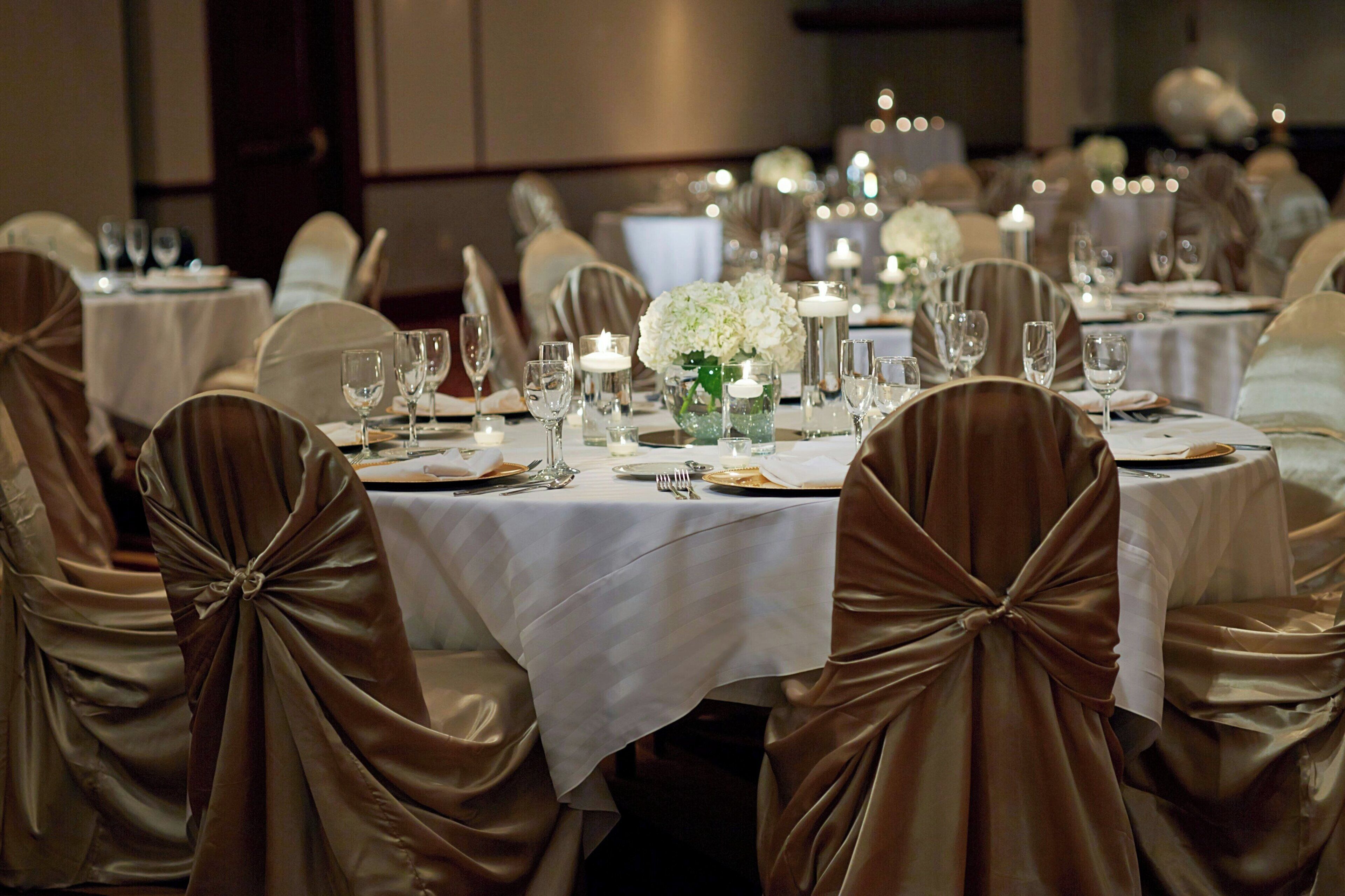 Un salón de banquetes con mesas redondas dispuestas para cenar, cada una con mantel blanco, cubertería y velas.
