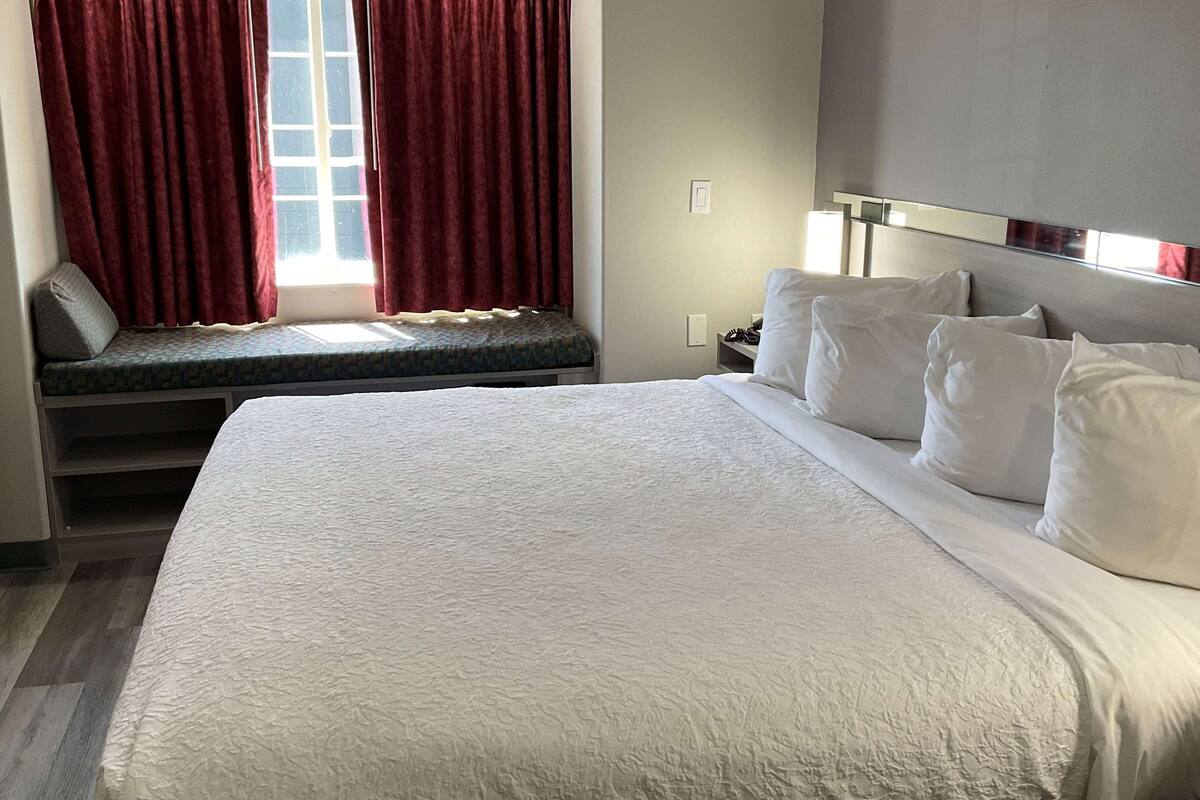 Ein Hotelzimmer mit einem großen Bett, einem Sitzfenster mit roten Vorhängen, einem Nachttisch mit Lampe und einem Kopfteil mit Kissen.