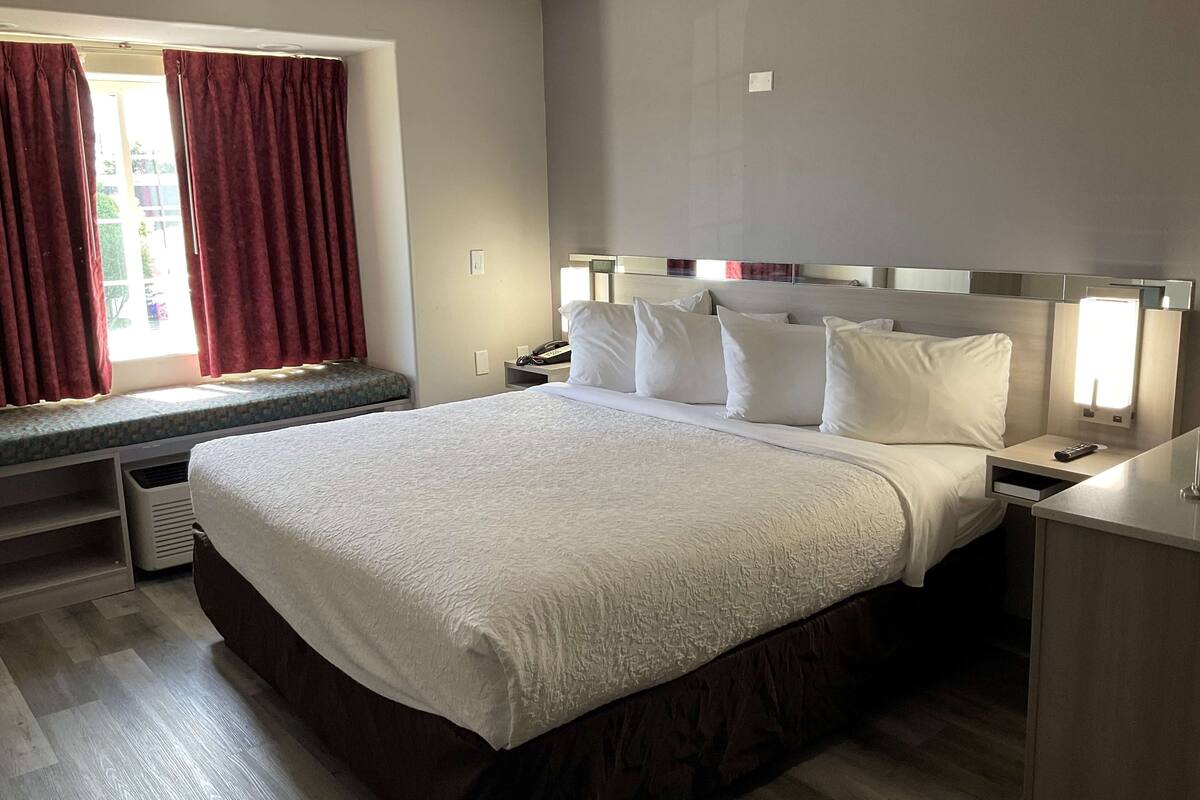 Ein Hotelzimmer mit einem großen Bett, einem Nachttisch mit Lampe, einem Fenster mit Vorhängen und Blick ins Freie.