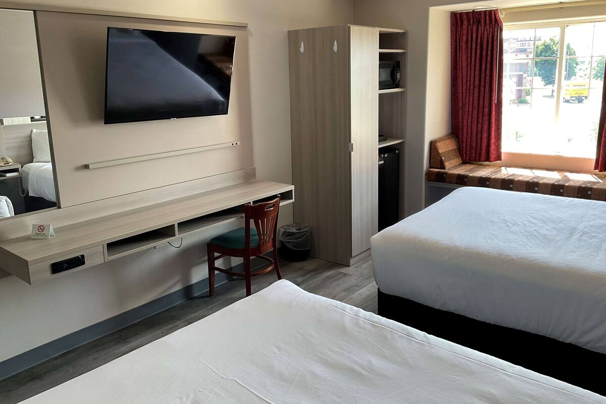Ein Hotelzimmer mit Bett, Schreibtisch mit Stuhl, Wandfernseher, Kleiderschrank und Fenster mit Blick nach draußen.