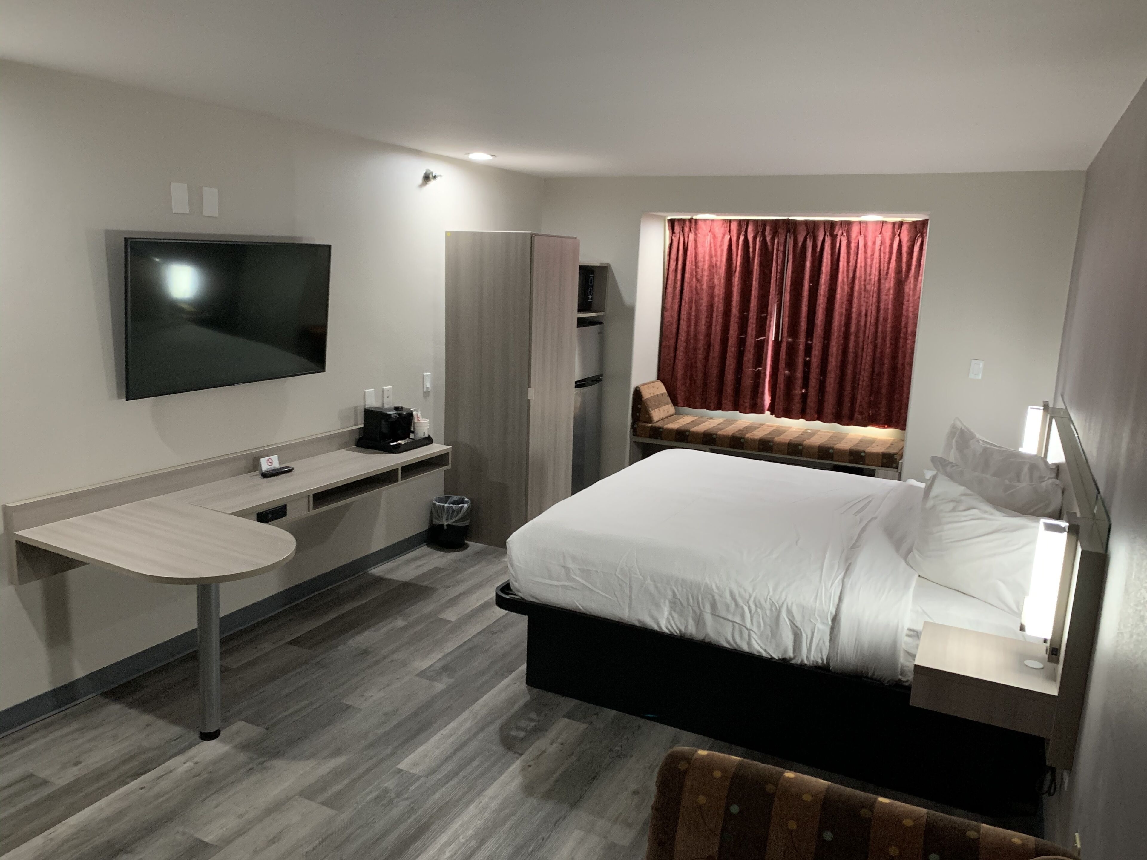 Una habitación de hotel con cama, escritorio, televisor, ventana con cortinas y un taburete pequeño.