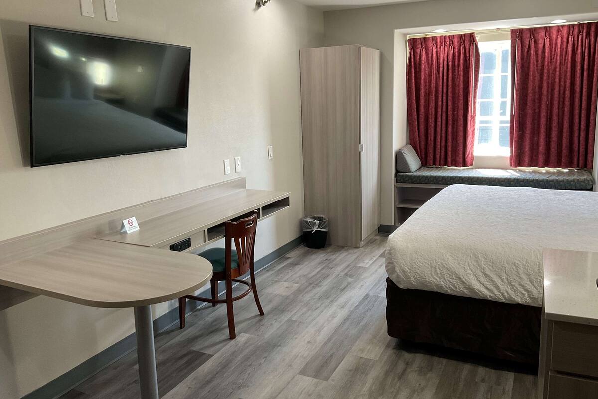 Ein Hotelzimmer mit Bett, Schreibtisch mit Stuhl, Wand-Fernseher und einem Fenster mit Vorhängen.