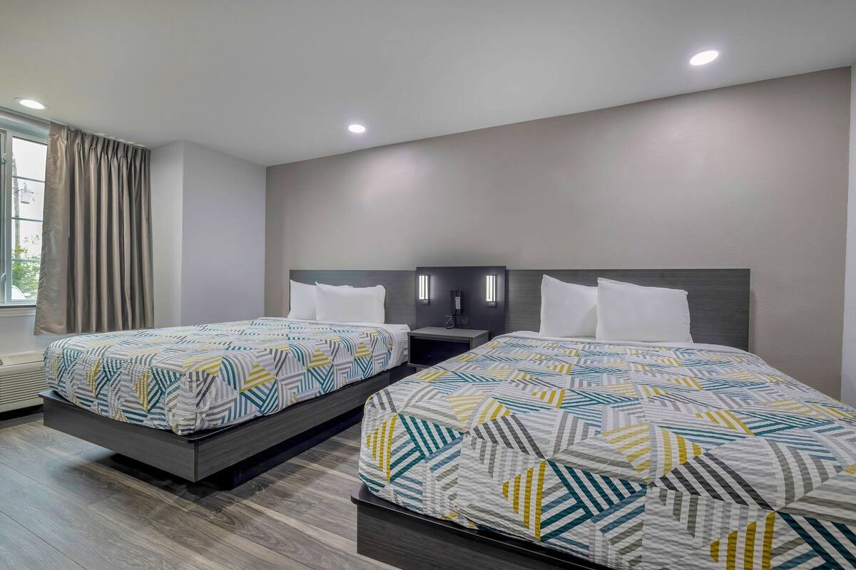 A hotel room with two beds, each with a patterned bedspread, a dark headboard, bedside tables with lamps, and a large window with curtains.
