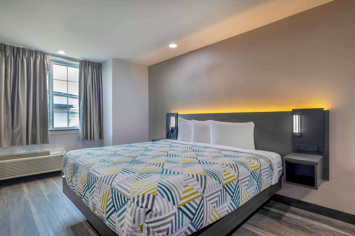 A hotel room with a bed, a nightstand, a wall-mounted lamp, and a window with curtains.