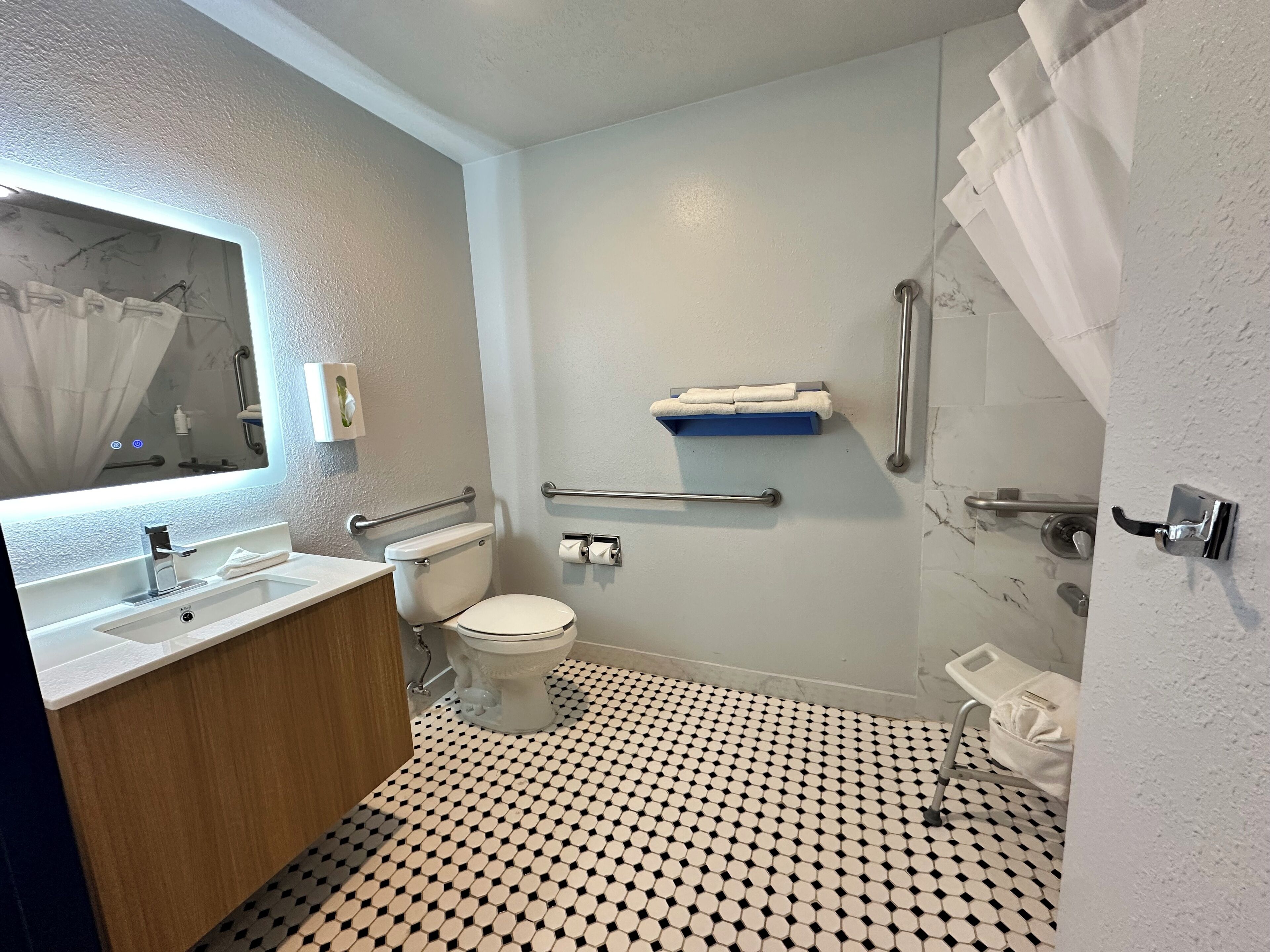 Un baño con inodoro blanco, lavamanos con espejo, zona de ducha con puerta de cristal y suelo estampado.