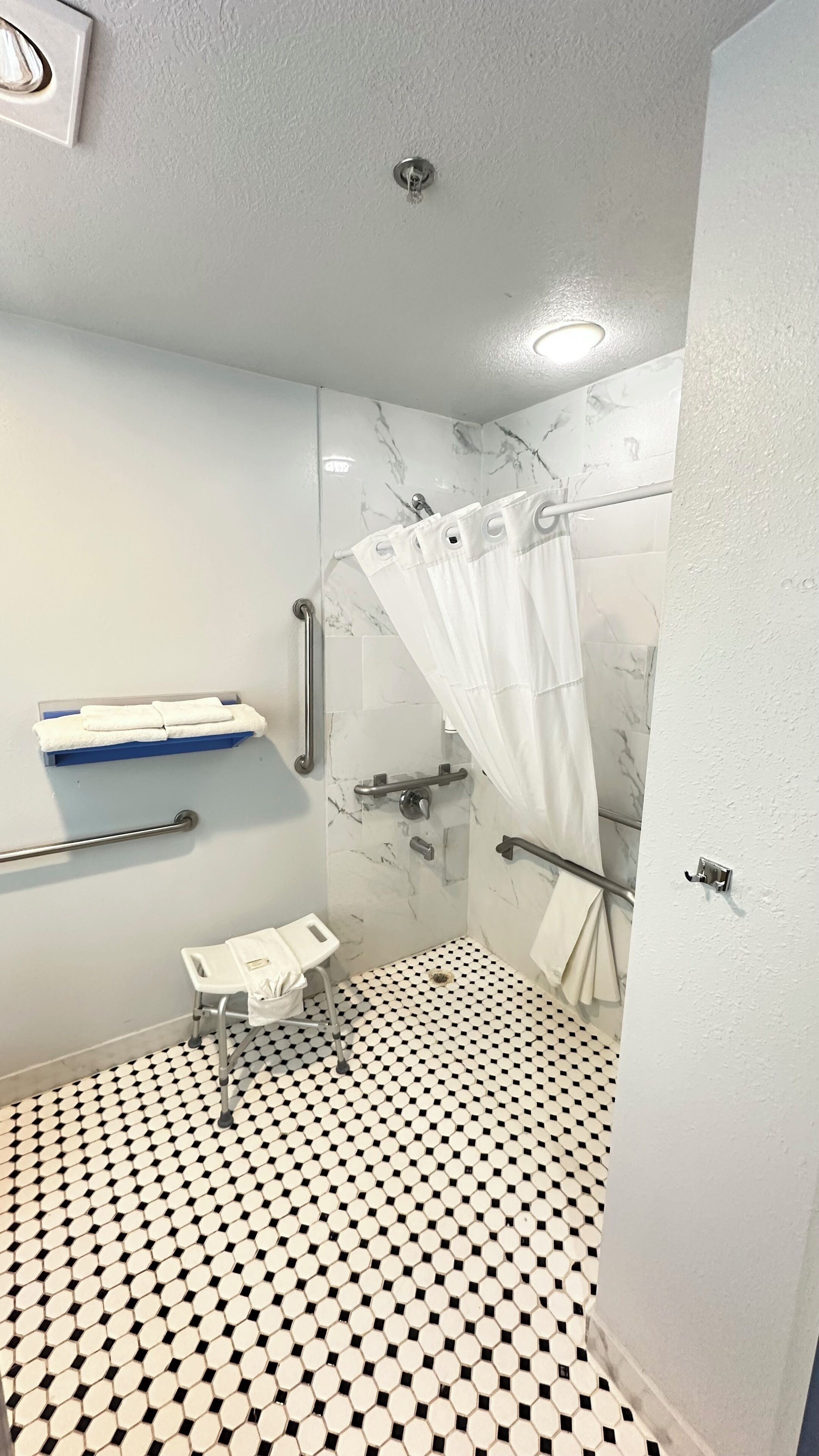 Un baño con piso a cuadros blanco y negro, una ducha con cortina blanca, un perchero y una barra de apoyo.