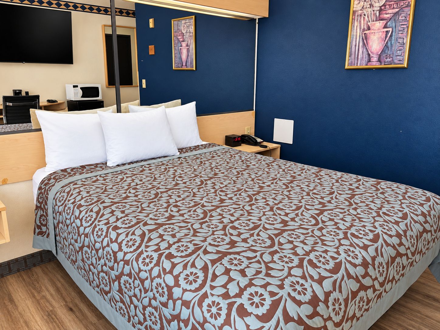 A hotel room with a large bed, a bedside table, a desk, a chair, a flat-screen TV, and artwork on the walls.