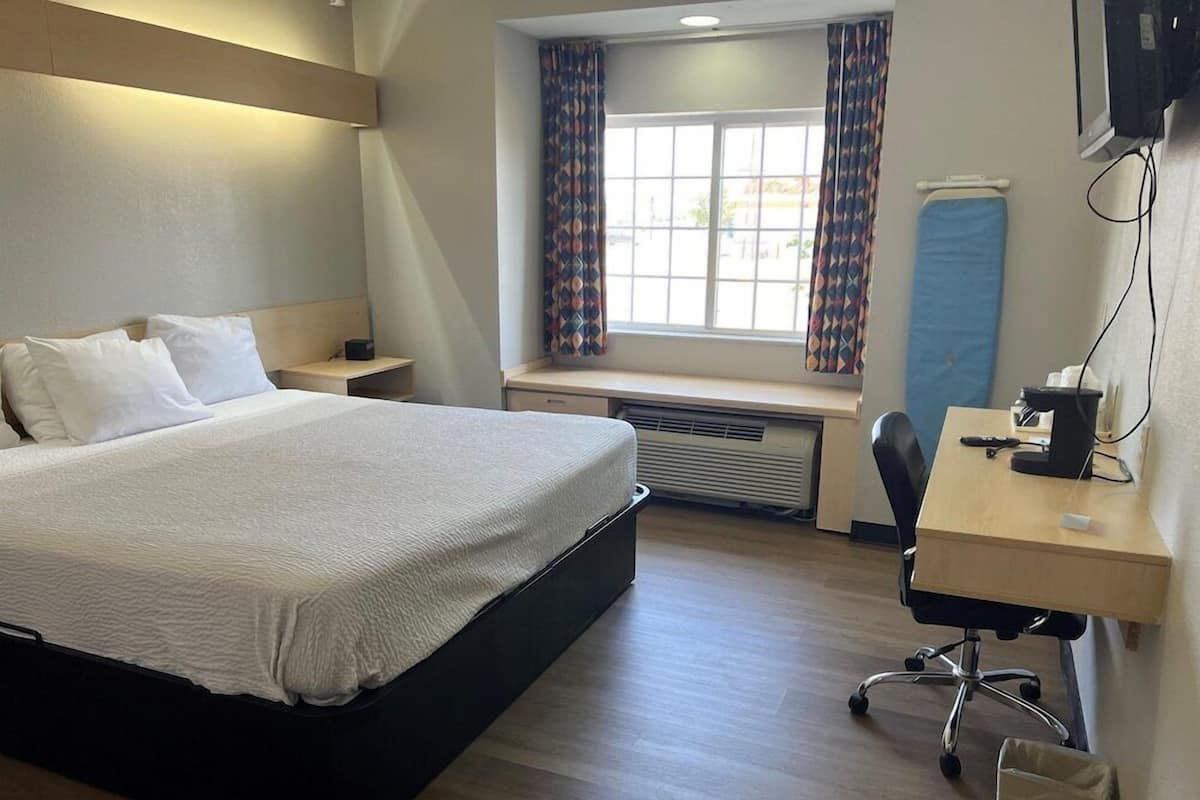 Een hotelkamer met een bed, een bureau, een stoel, een raam met gordijnen, een verwarming en een aan de muur bevestigde televisie.