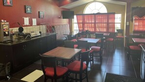 A restaurant interior with red chairs, wooden tables, a counter with a waffle maker, and a large window with curtains.