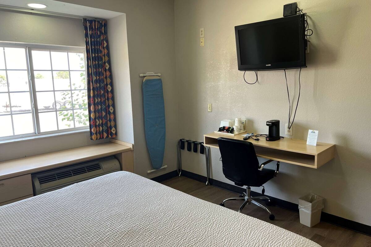 Een hotelkamer met een bed, een bureau met stoel, een aan de muur bevestigde televisie en een raam met gordijnen.