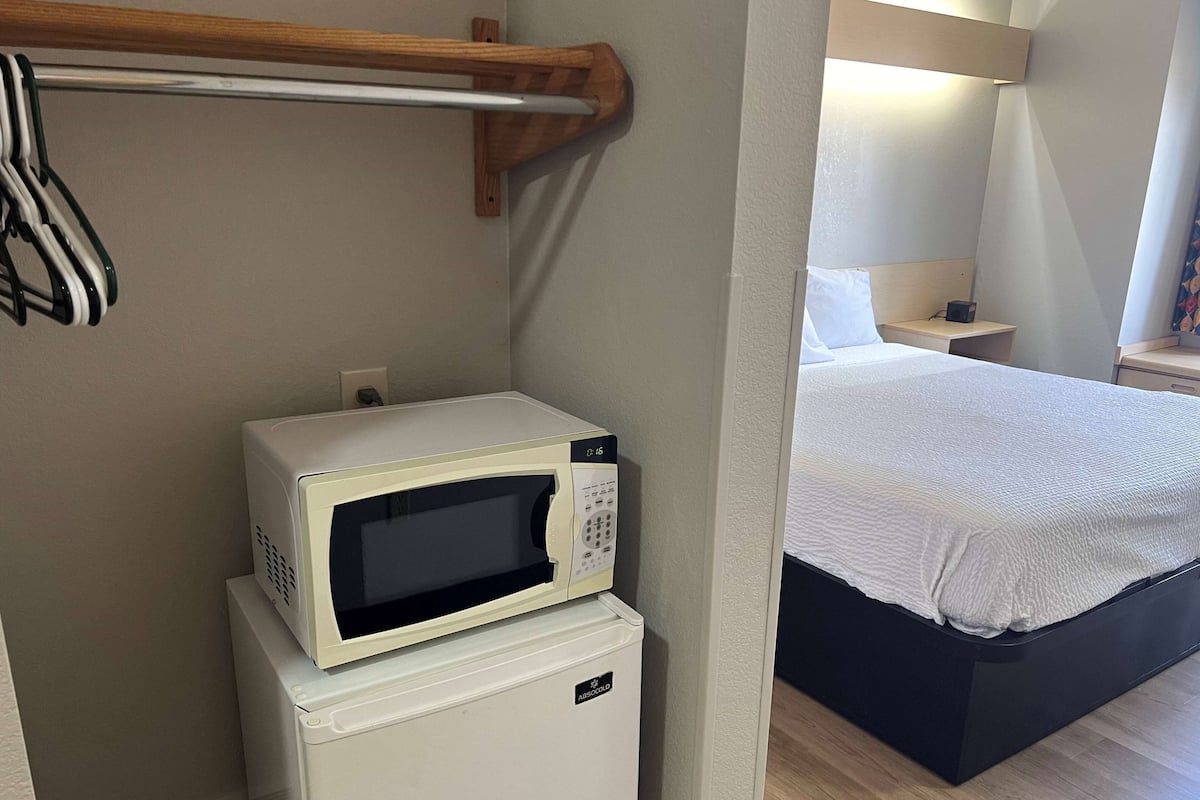 Een kleine kamer met een bed, een nachtkastje, een magnetron en een koelkast.