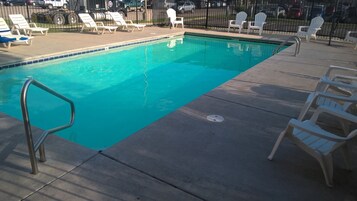 Una piscina con una escalera metálica, rodeada de sillas reclinables blancas y una cubierta de madera.
