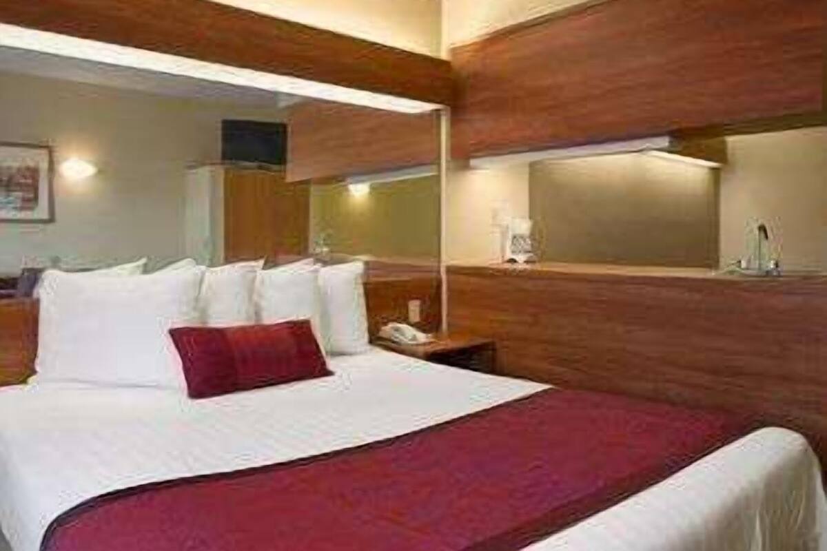 A hotel room with a large bed, wooden paneling, a nightstand with a lamp, and a mirror above the bed.