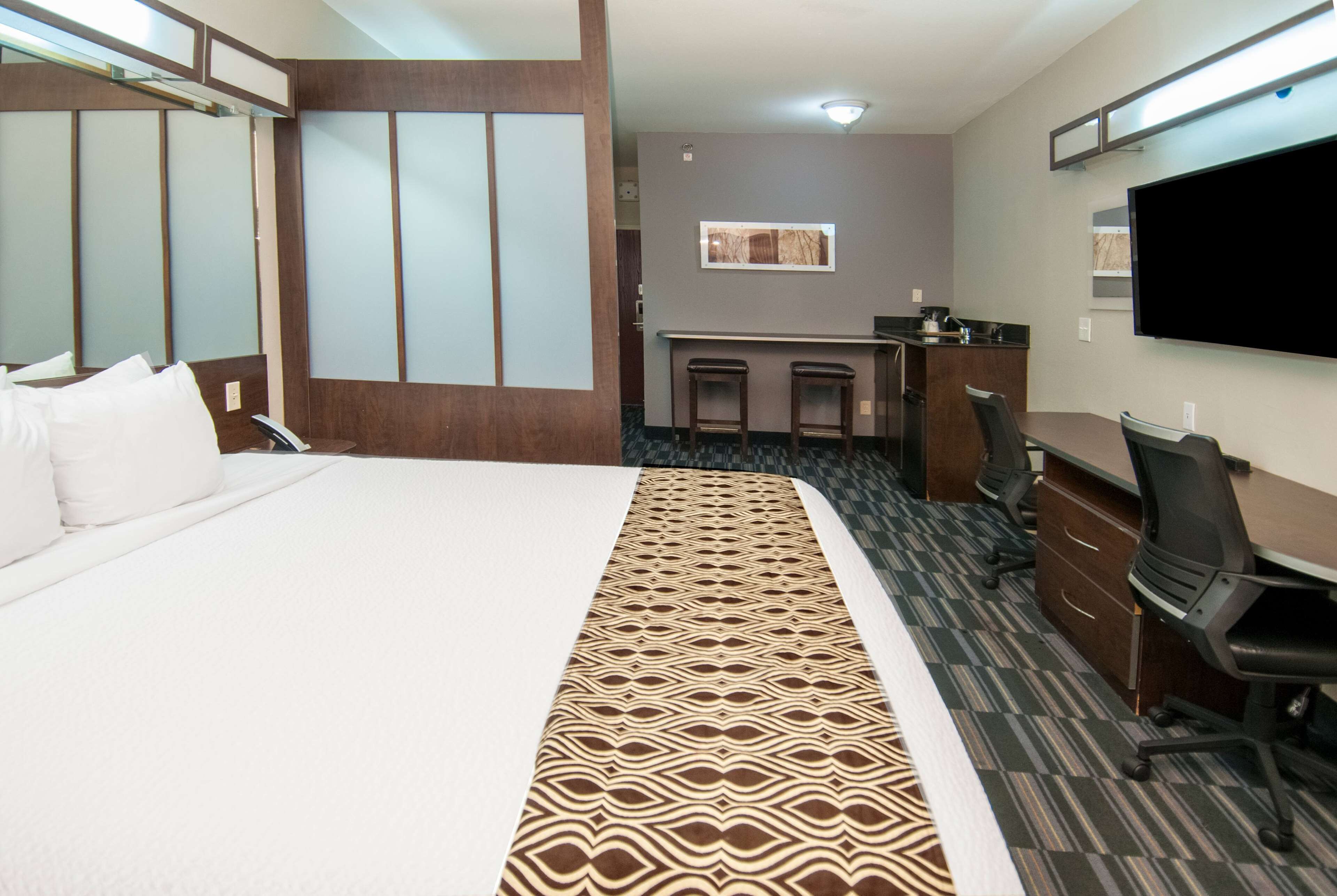 A hotel room with a large bed, a desk with two chairs, a flat-screen TV, and a small kitchenette.