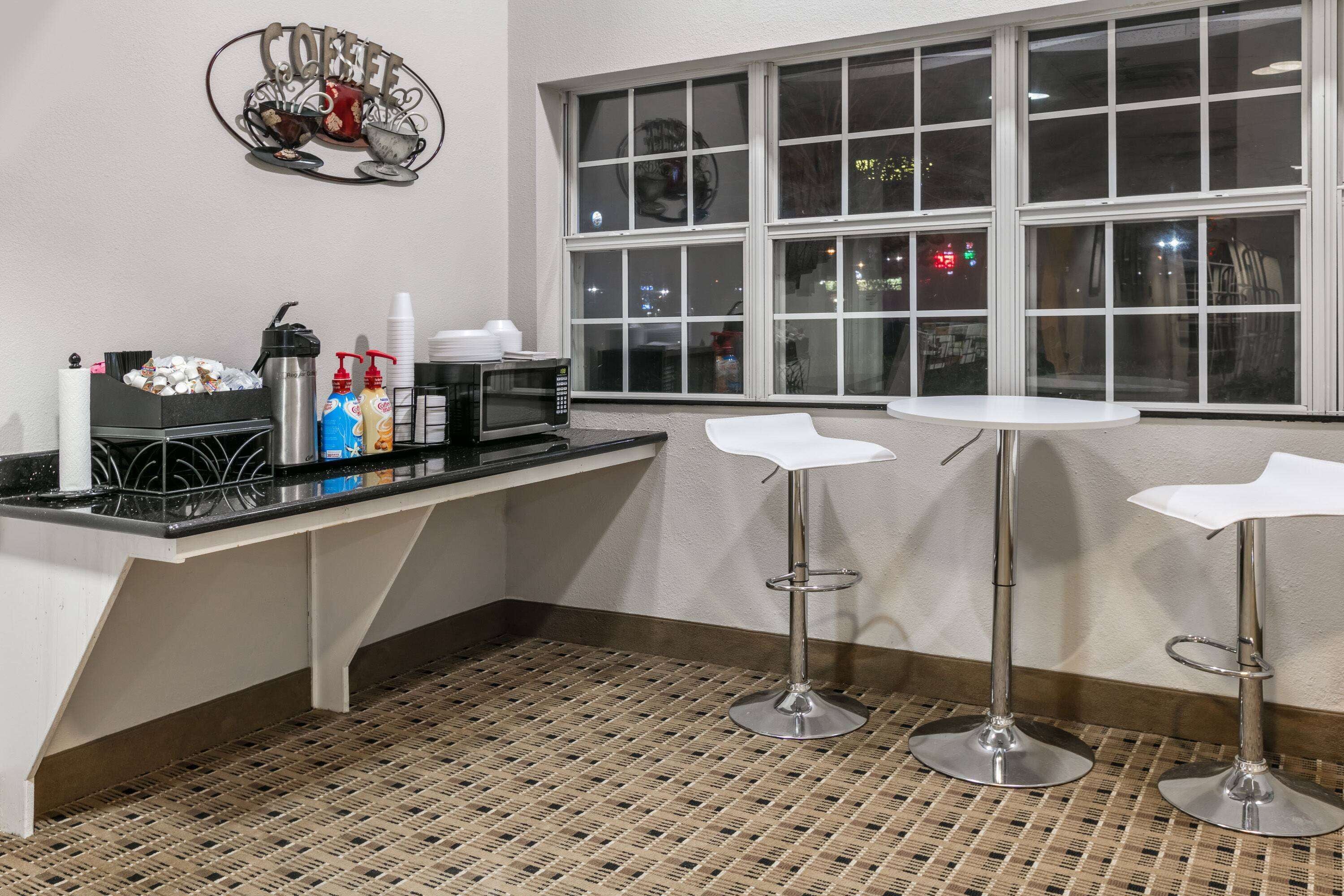 Una zona de cocina con microondas, cafetera y estación para café. Hay dos taburetes de bar y una ventana con vista a la ciudad de noche.