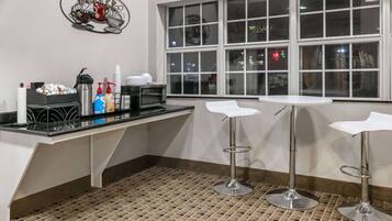 Una zona de cocina con microondas, cafetera y estación para café. Hay dos taburetes de bar y una ventana con vista a la ciudad de noche.
