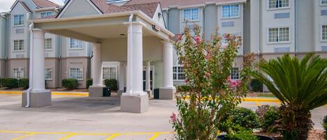 A multi-story hotel with a covered entrance, a parking lot, and landscaped areas with potted plants.