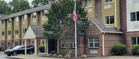 A multi-story hotel with a brick facade, a flagpole with an American flag, and a parking area with cars.