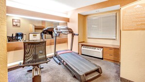 A hotel room with a treadmill, a desk, a chair, and a large window with blinds. - Microtel Inn by Wyndham Newport News Airport (Newport News)