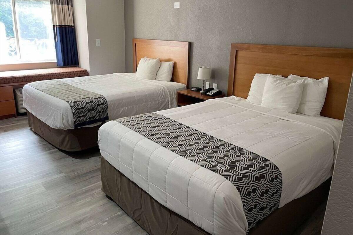 A hotel room with two beds, each with a wooden headboard, white bedding, and a patterned throw. There is a window with curtains, a bedside table with a lamp, and a small shelf.