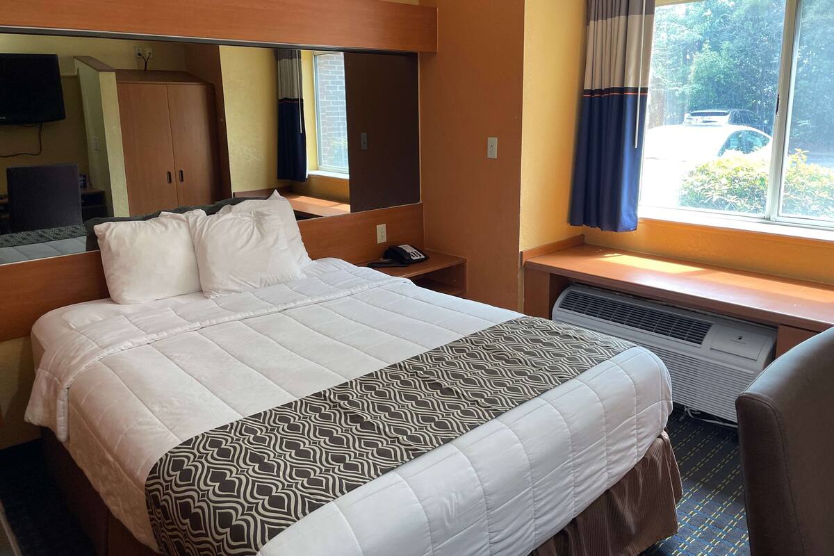 A hotel room with a bed, a desk with a computer, a mirror, a wardrobe, and a window with curtains.