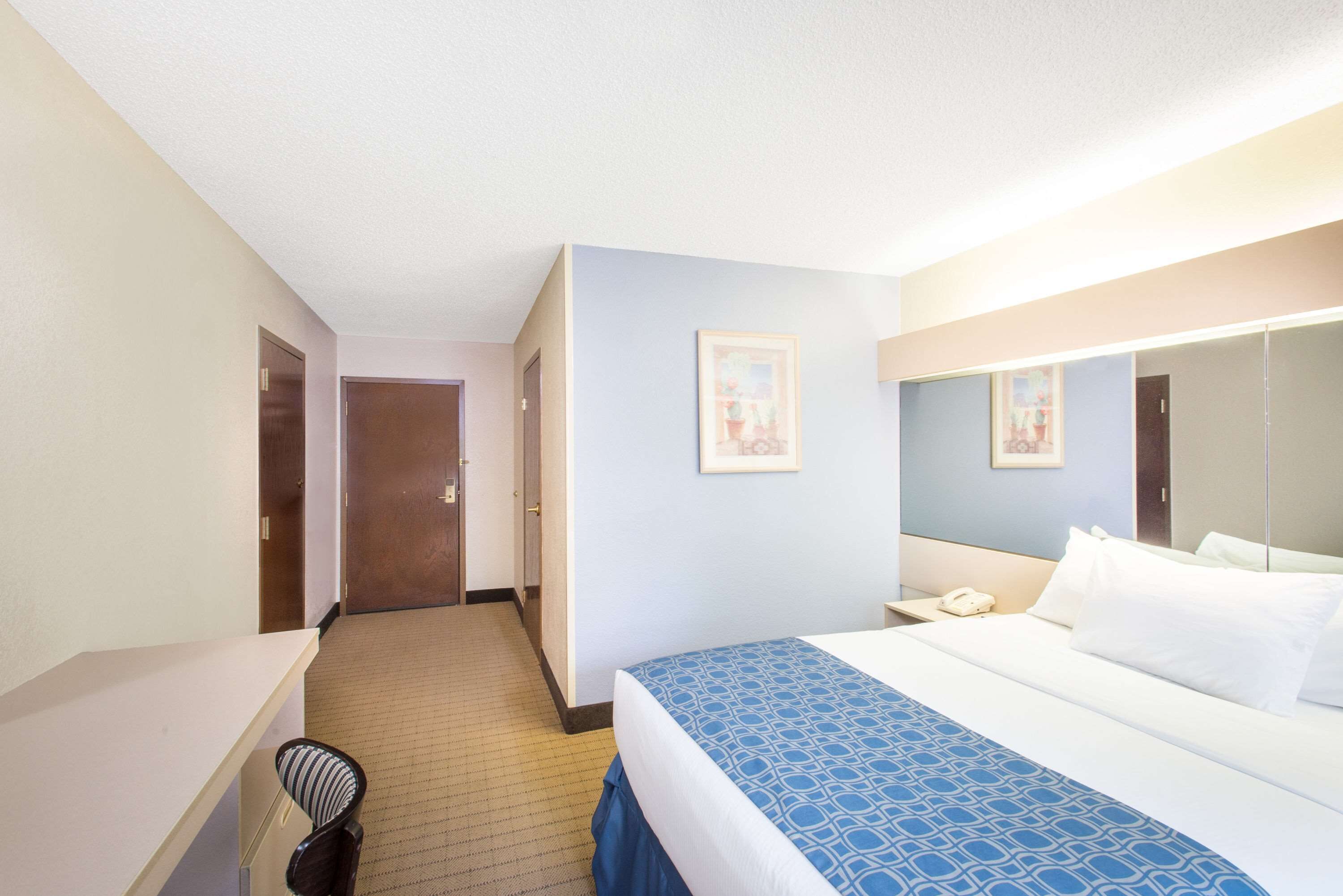 A hotel room with a bed, bedside table, and a mirror. There is a door leading to another room and a hallway.