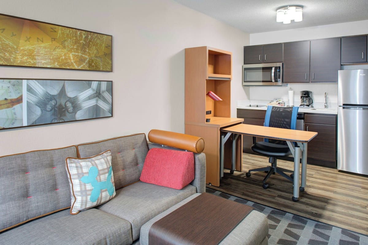 A compact living space with a sofa, a small desk, a chair, a kitchenette, and artwork on the walls.