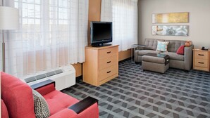 A hotel room with a TV on a wooden stand, a sofa, a chair, a small table, and a heater. - TownePlace Suites by Marriott Wichita East (Wichita)