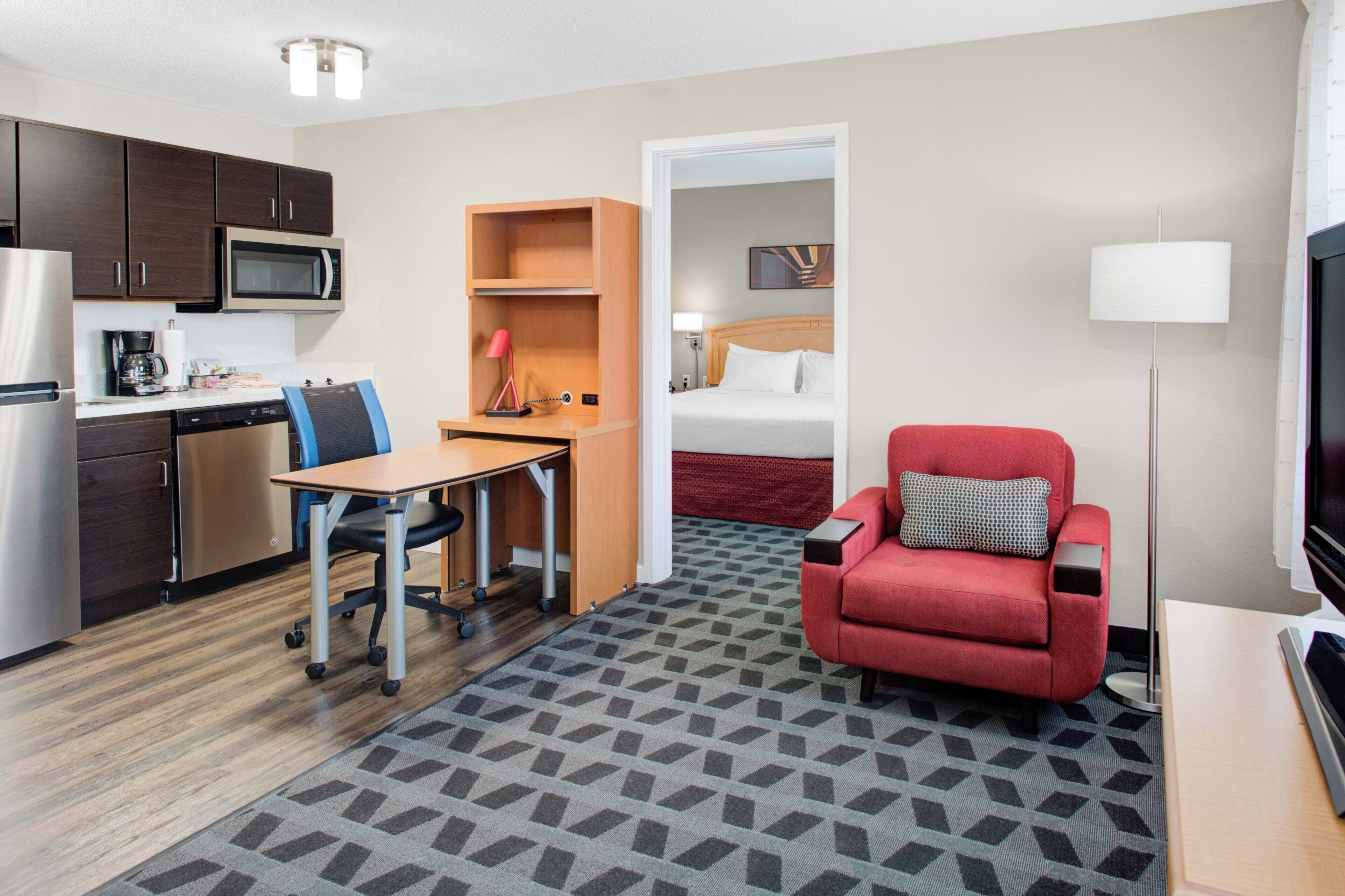 A hotel room with a kitchenette, a desk with a chair, a red sofa, a bed, and a TV.
