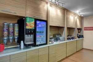Un área de desayuno con dispensador de bebidas autoservicio, un refrigerador lleno de bebidas y una cafetera.