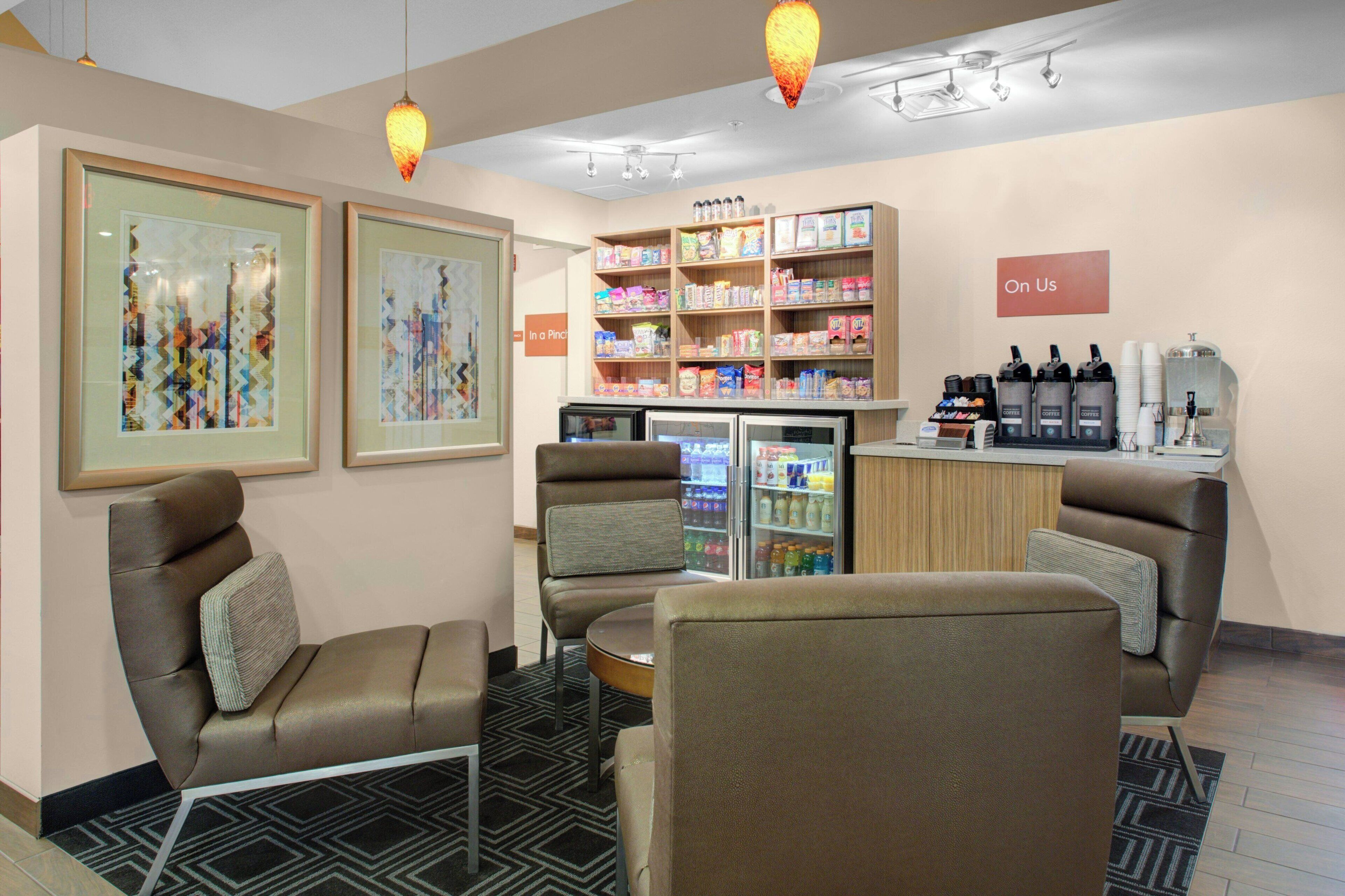 A hotel lobby with seating, a refrigerator stocked with beverages, and a sign reading "On Us".