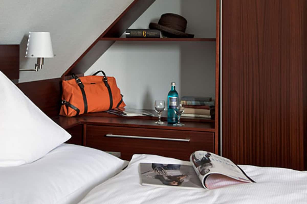 A hotel room with a bed, a nightstand, a wooden wardrobe, a lamp, a suitcase, a bottle, and a magazine.