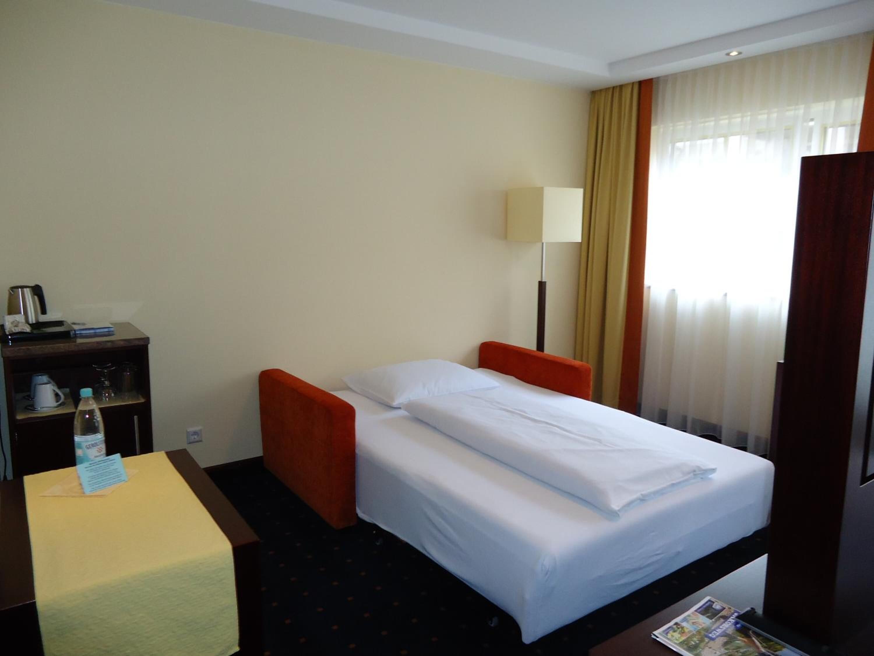Quarto de hotel com cama, escrivaninha com luminária e janela com cortinas.