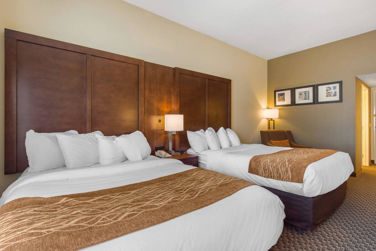 A hotel room with two beds, a large wooden headboard, a nightstand with a lamp, and framed pictures on the wall.