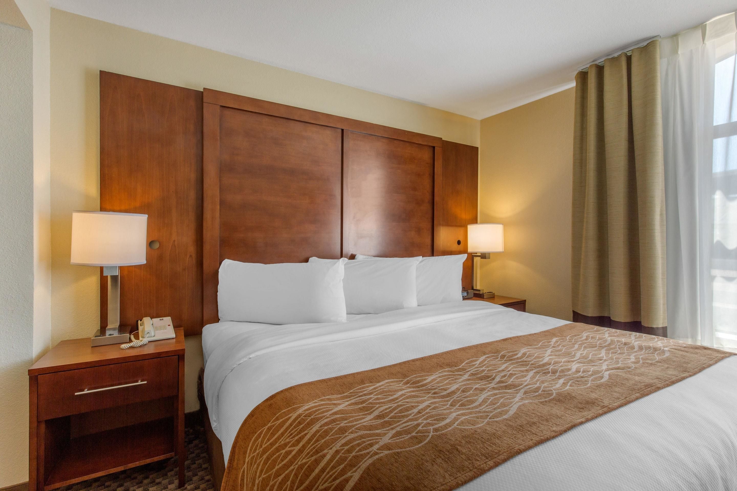 Une chambre d’hôtel avec un grand lit, une tête de lit en bois, deux tables de chevet avec des lampes et une fenêtre avec des rideaux.