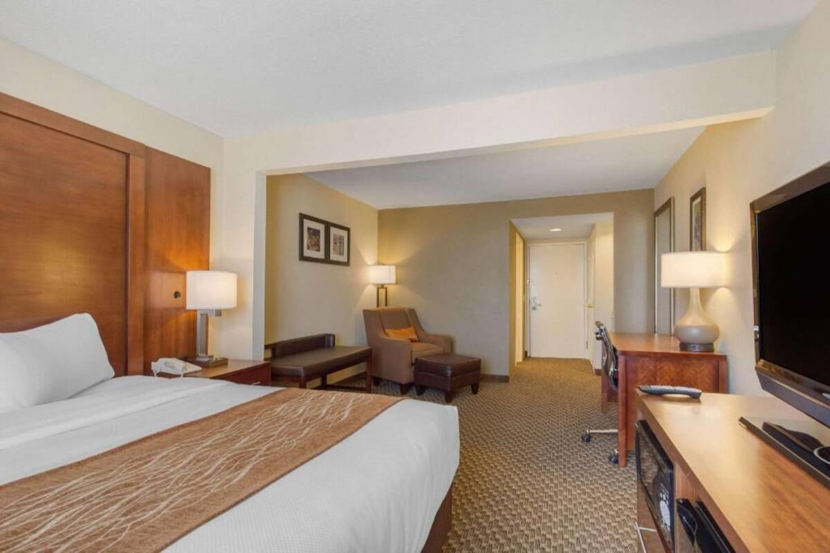 A hotel room with a large bed, a desk with a TV, a chair, a lamp, and a door leading to another room.