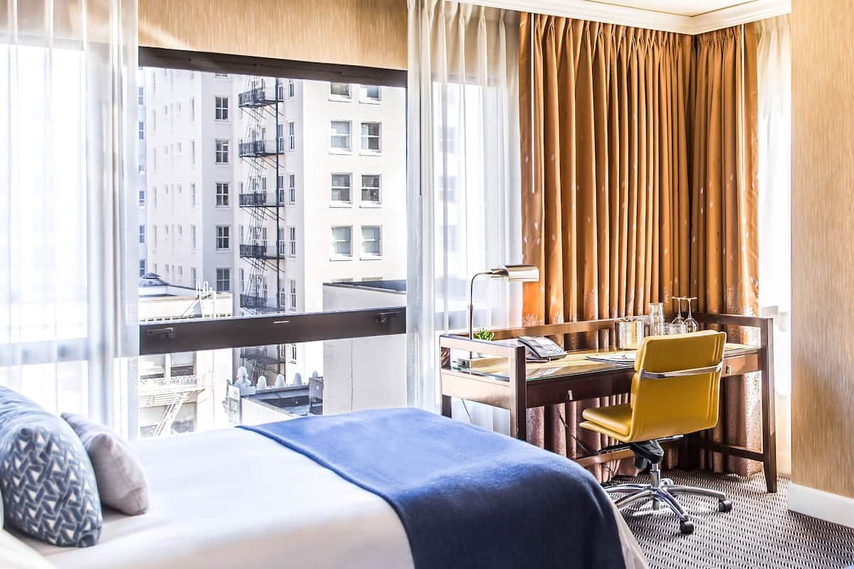 A hotel room with a bed, a desk with a chair, a lamp, and a view of the city through large windows.