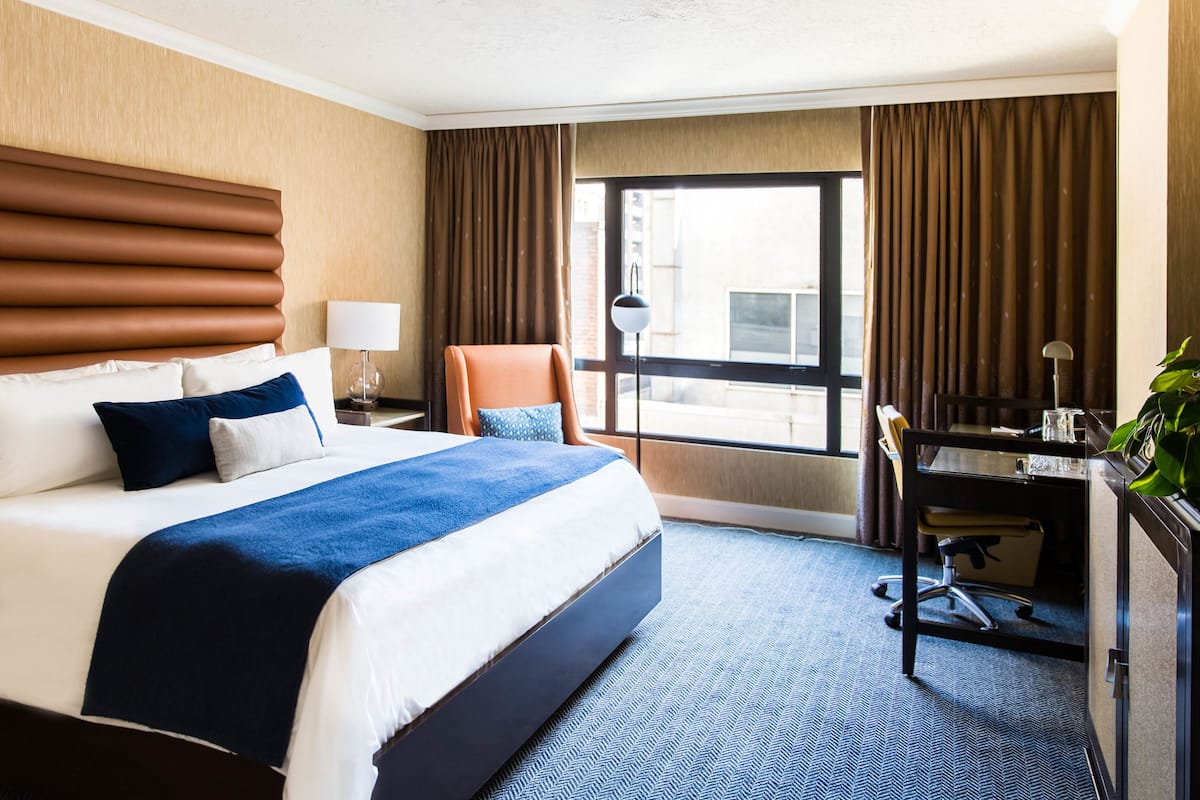 A hotel room with a large bed, a desk, a chair, and a view of the city through the window.